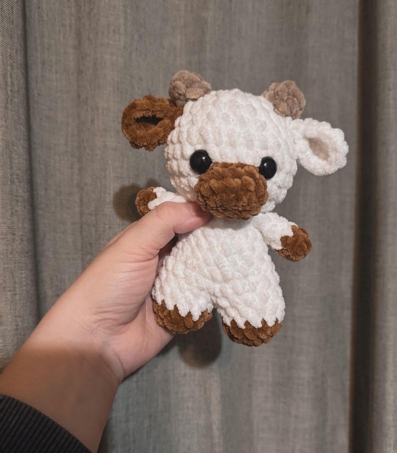 May include: A white crocheted stuffed animal shaped like a cow with brown crocheted ears, nose, and hooves. The cow has black button eyes.