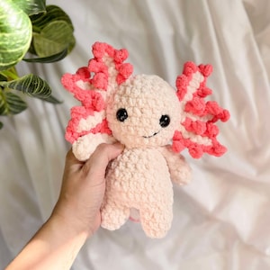 May include: A crocheted axolotl plush toy with pink gills and black eyes. The toy is made of soft, fluffy yarn and is about 6 inches tall.
