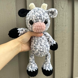 May include: A crocheted stuffed cow toy with black and white speckled fur, black hooves, and a pink nose. The cow has yellow horns and black eyes.