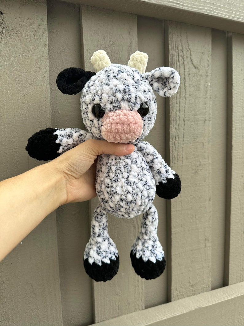 May include: A crocheted stuffed cow toy with black and white speckled fur, black hooves, and a pink nose. The cow has yellow horns and black eyes.