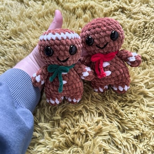 May include: Two brown crocheted gingerbread man plushies with white icing details and smiling faces. One gingerbread man has a green bow and the other has a red bow.
