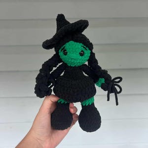 May include: A handmade crochet doll of a witch with a green face, black hat, dress, and braids. The doll has large black feet and is held in a person's hand. The witch doll is a whimsical, handcrafted item.