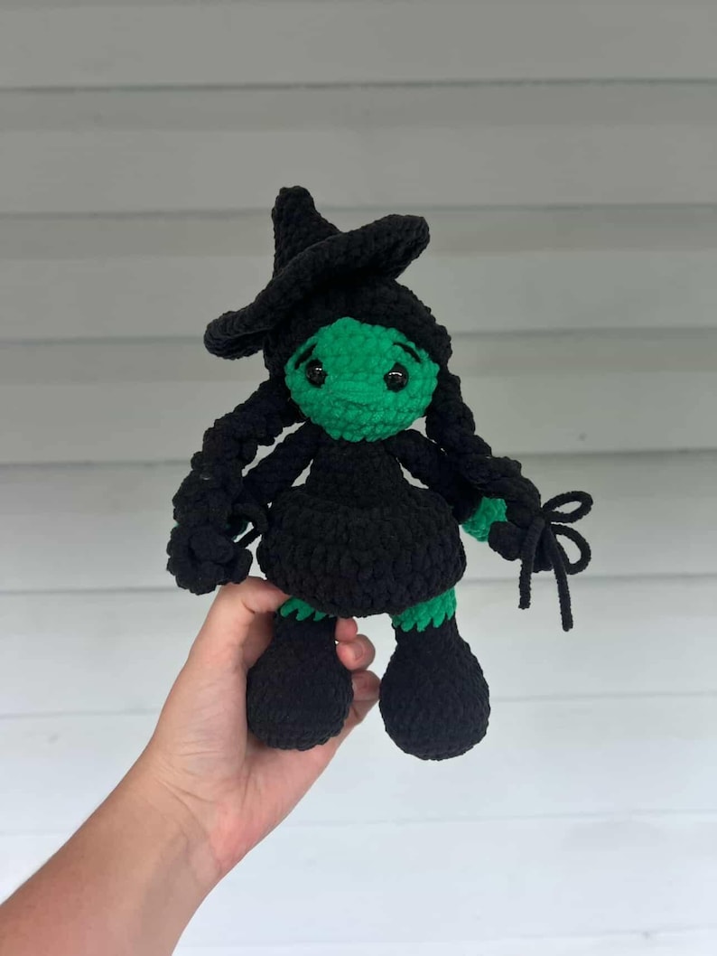 May include: A handmade crochet doll of a witch with a green face, black hat, dress, and braids. The doll has large black feet and is held in a person's hand. The witch doll is a whimsical, handcrafted item.