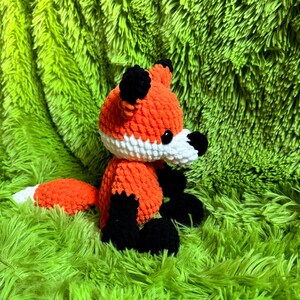 May include: A crocheted orange and black fox plush toy with white accents. The toy is sitting on a green fuzzy surface.