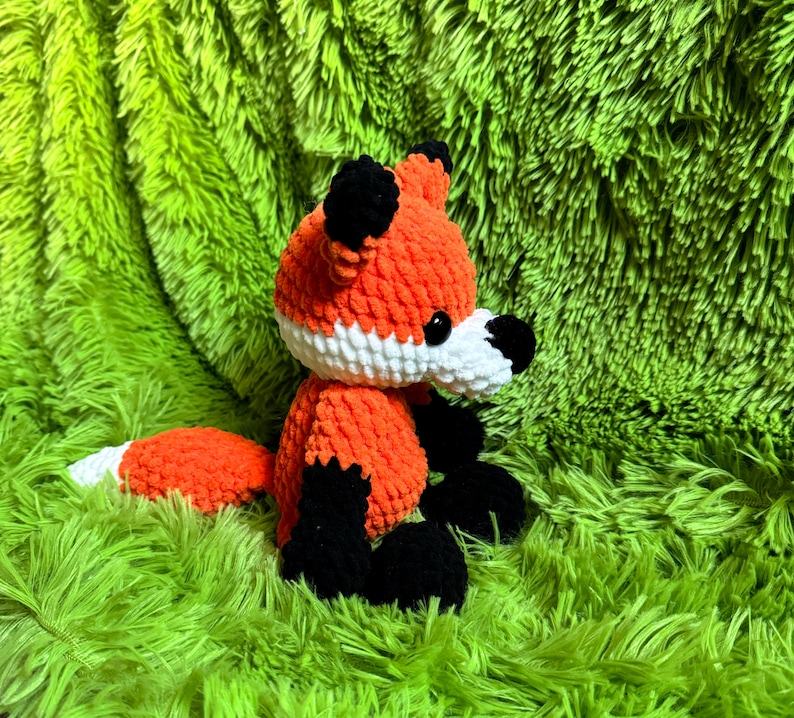 May include: A crocheted orange and black fox plush toy with white accents. The toy is sitting on a green fuzzy surface.