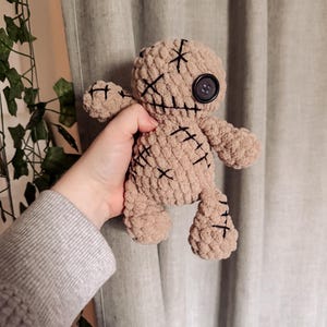 Könnte beinhalten: Eine beige Häkelpuppe mit schwarzen Nahtdetails und einem großen schwarzen Knopf als Auge. Die Puppe hat aufgenähte Kreuzmarkierungen auf Gesicht und Körper. Die Puppe wird in einer Hand gehalten.