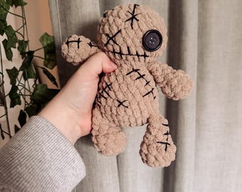 May include: A tan crochet doll with black stitched details and a large black button eye. The doll has stitched cross marks on its face and body. The doll is held in a hand against a neutral background.