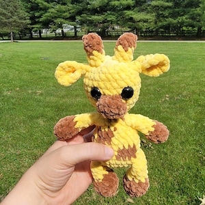 May include: A handmade crochet giraffe plush toy. The giraffe is yellow with brown spots, ears, and hooves. It has large black safety eyes and is being held in front of a green grassy background.