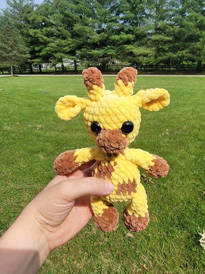 May include: A handmade crochet giraffe plush toy. The giraffe is yellow with brown spots, ears, and hooves. It has large black safety eyes and is being held in front of a green grassy background.