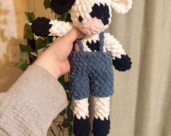 May include: A handmade crochet cow doll with black and white spots, black hooves, and blue overalls. The doll has small brown horns and pink cheeks. The arms and legs are outstretched, giving the appearance of a friendly character.