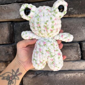 以下が含まれることがあります： ピンクとグリーンの花柄の白い毛糸で編まれたカエルのぬいぐるみ。カエルは大きな黒い目と小さく丸い体をしている。