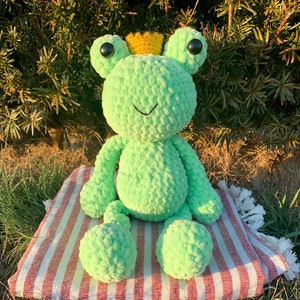 May include: A green crocheted frog wearing a yellow crown. The frog is sitting on a red and white striped surface.