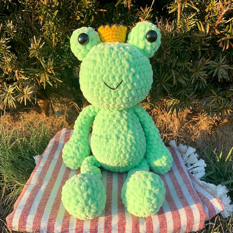 May include: A green crocheted frog wearing a yellow crown. The frog is sitting on a red and white striped surface.