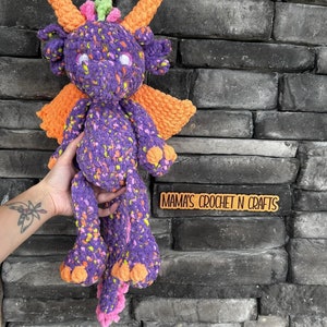 May include: Purple and orange crocheted dragon toy with a speckled pattern. The dragon has a long tail, large wings, and a friendly face. The toy is held in a hand against a brick wall. The text "MAMA'S CROCHET N CRAFTS" is visible on a wooden sign.