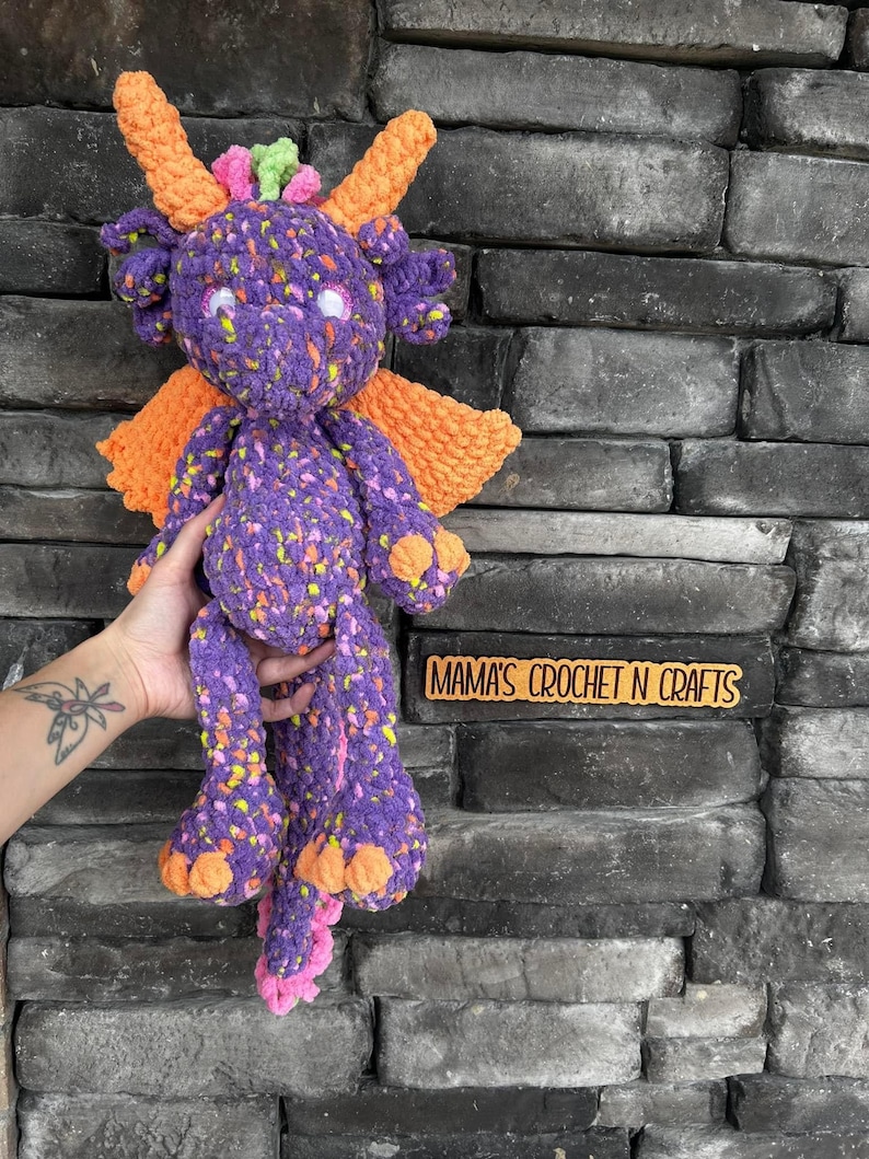May include: Purple and orange crocheted dragon toy with a speckled pattern. The dragon has a long tail, large wings, and a friendly face. The toy is held in a hand against a brick wall. The text "MAMA'S CROCHET N CRAFTS" is visible on a wooden sign.