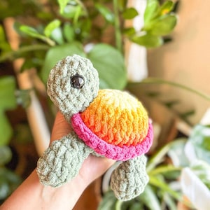 May include: A handmade crochet turtle toy. The turtle has a green body, a shell with orange, yellow, and pink colors, and a black eye. The toy is held in a person's hand, with green plants in the background.