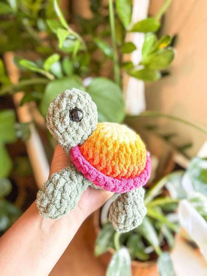 May include: A handmade crochet turtle toy. The turtle has a green body, a shell with orange, yellow, and pink colors, and a black eye. The toy is held in a person's hand, with green plants in the background.
