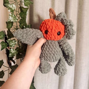 May include: A handmade crochet plush toy in the shape of a creature. The toy has a round orange head with black eyes and small brown ears. The body, wings, and feet are gray. The toy is held by a hand.