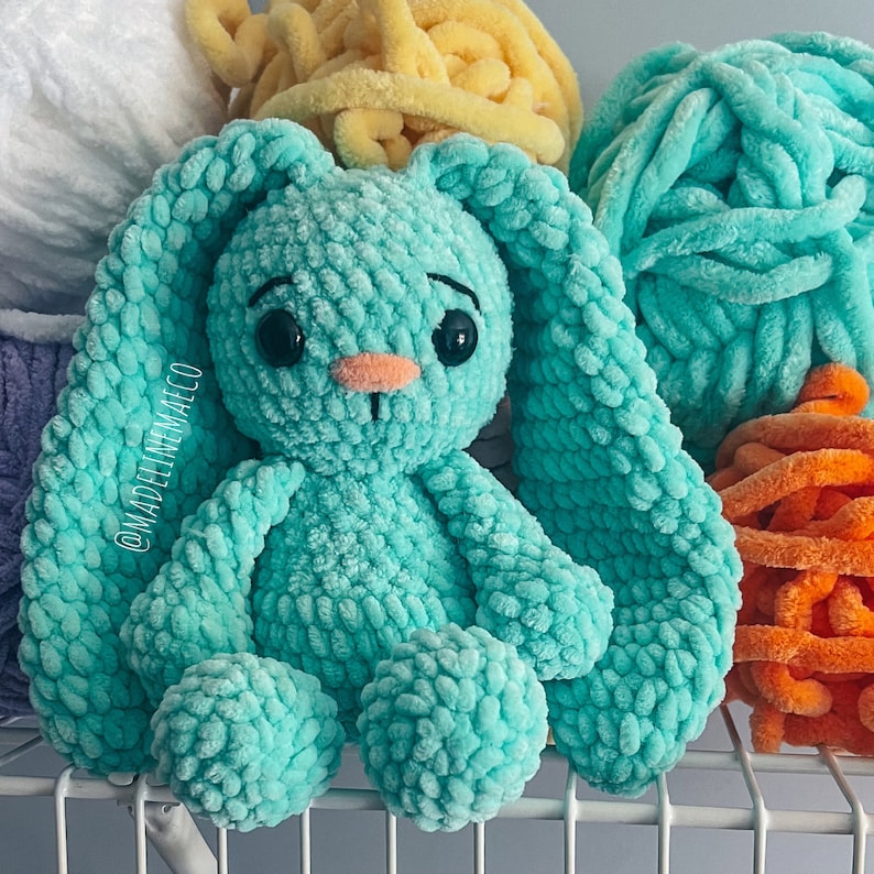 May include: A teal blue crocheted bunny rabbit toy with a pink nose and black eyes. The bunny is sitting on a white shelf with colorful yarn in the background. The text '@MadelineMaeco' is visible on the bunny's left ear.