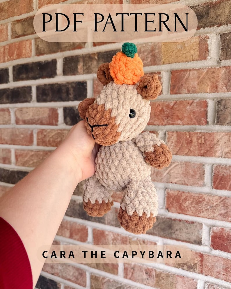 May include: A crochet capybara plush toy with light tan and brown yarn, featuring a small orange pumpkin accessory on its head. The text PDF PATTERN and CARA THE CAPYBARA are visible on the image, which depicts a soft, textured stuffed animal.