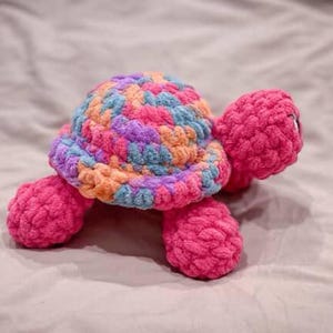 May include: A handmade crochet turtle in vibrant colors. The turtle's shell is a mix of blue, orange, purple, and pink yarn, while the head, legs, and tail are a solid bright pink. The plush toy is soft and cuddly.