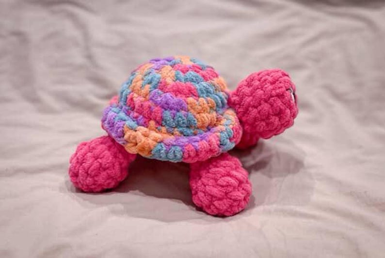 May include: A handmade crochet turtle in vibrant colors. The turtle's shell is a mix of blue, orange, purple, and pink yarn, while the head, legs, and tail are a solid bright pink. The plush toy is soft and cuddly.