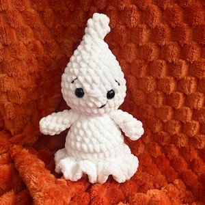 Puede incluir: Un juguete de peluche de fantasma tejido a ganchillo en color blanco con una parte superior puntiaguda, pequeños ojos de plástico negro y una boca cosida. La figura tiene una cabeza redondeada, dos brazos pequeños y una base con volantes.