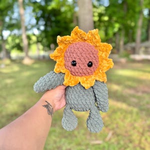 K&ouml;nnte beinhalten: Ein geh&auml;keltes Pl&uuml;sch-Sonnenblumenspielzeug mit einem grauen K&ouml;rper und einem gelben und rosafarbenen Sonnenblumenkopf. Das Spielzeug hat schwarze Augen und einen freundlichen Ausdruck.
