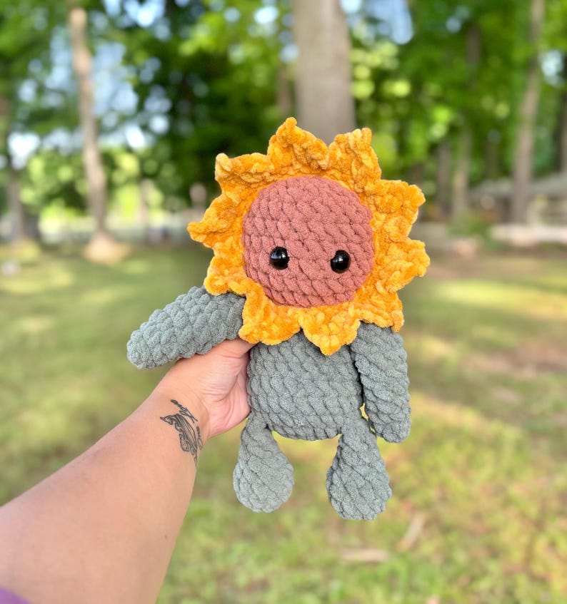 K&ouml;nnte beinhalten: Ein geh&auml;keltes Pl&uuml;sch-Sonnenblumenspielzeug mit einem grauen K&ouml;rper und einem gelben und rosafarbenen Sonnenblumenkopf. Das Spielzeug hat schwarze Augen und einen freundlichen Ausdruck.