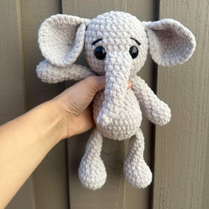 May include: A gray crocheted elephant plush toy with black eyes. The elephant is being held by a hand.