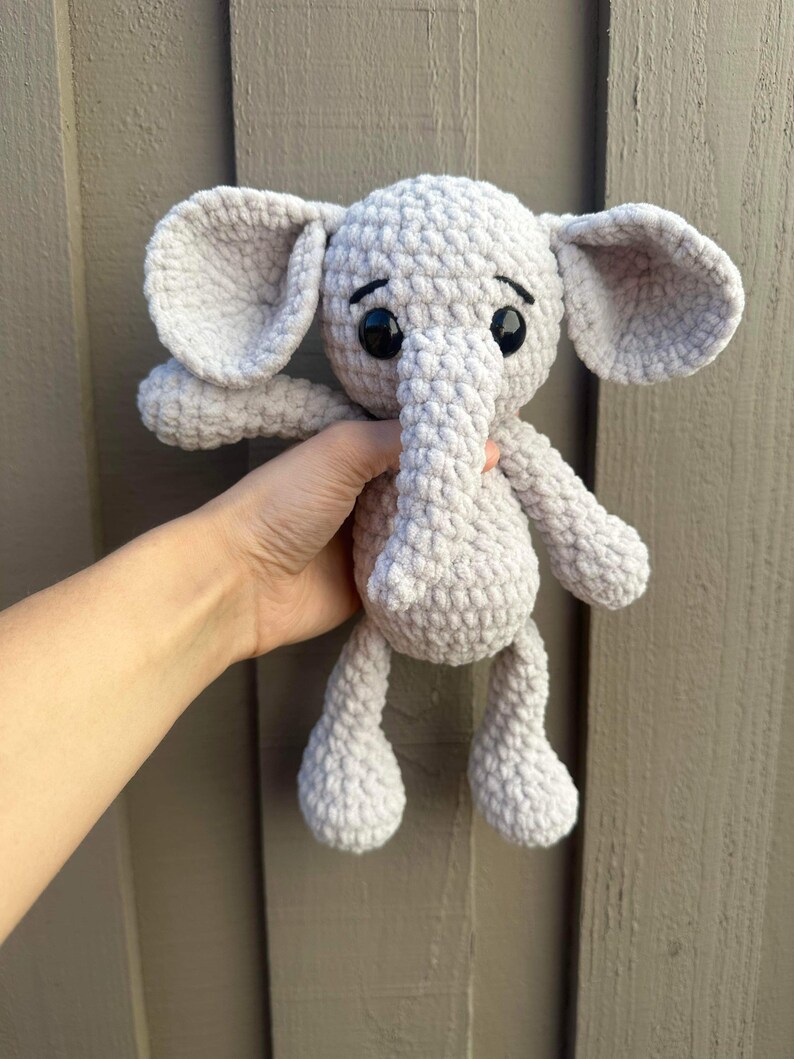 May include: A gray crocheted elephant plush toy with black eyes. The elephant is being held by a hand.