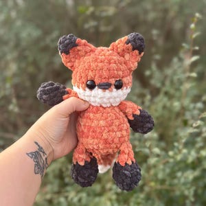 May include: A crocheted fox toy in orange, white, and grey. The fox has black eyes, a white muzzle, and grey paws and ear tips. The plushie is held in a hand against a blurred green backdrop.