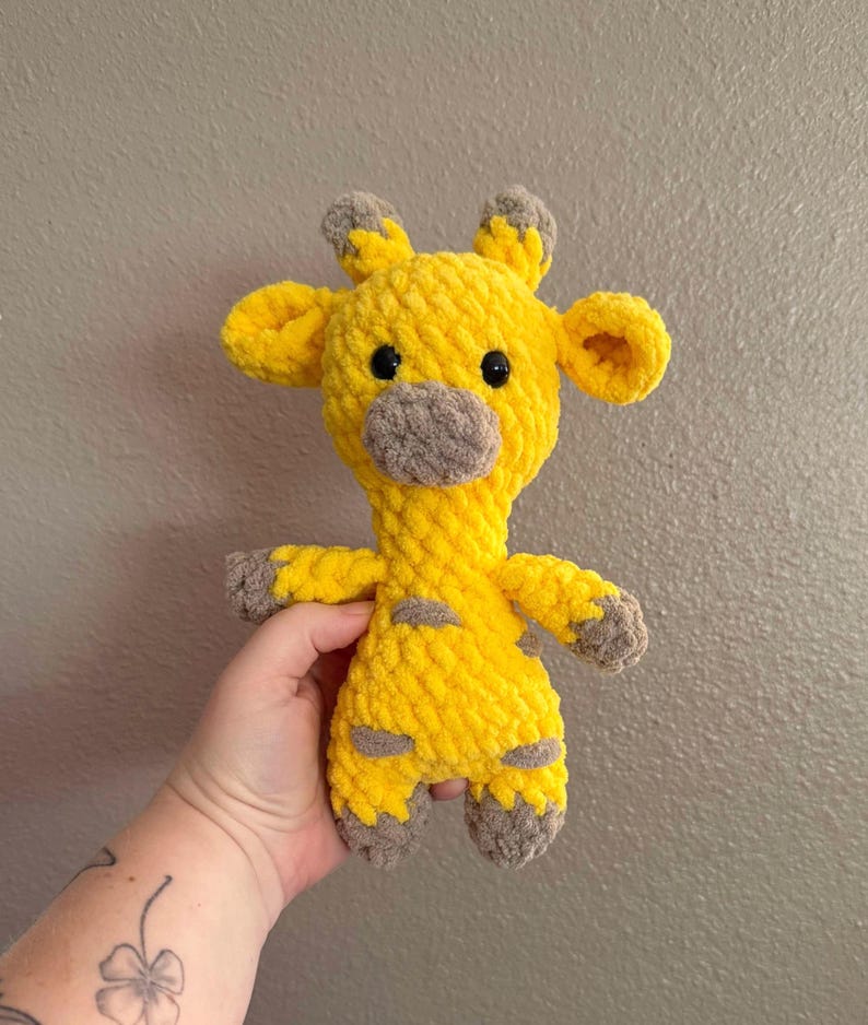 May include: A yellow crocheted giraffe stuffed animal with brown ears, nose, and feet. The giraffe has black eyes and a textured, fuzzy surface.
