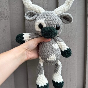 May include: A handmade crochet plush toy in the shape of a reindeer. The reindeer has a gray body with white and dark green accents. It features large black eyes and white antlers. The toy is held against a gray wooden background.