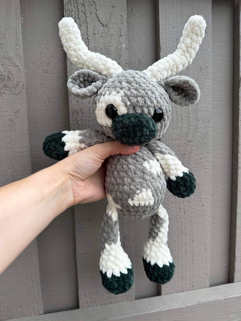 May include: A handmade crochet plush toy in the shape of a reindeer. The reindeer has a gray body with white and dark green accents. It features large black eyes and white antlers. The toy is held against a gray wooden background.