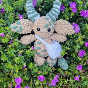 Peut inclure: Une peluche crochet&eacute;e &agrave; la main au design fantaisiste. Le jouet pr&eacute;sente un corps beige avec des taches vertes, de grandes oreilles et des cornes ray&eacute;es. Il a une sacoche blanche et est entour&eacute; de feuillage vert et de fleurs violettes.