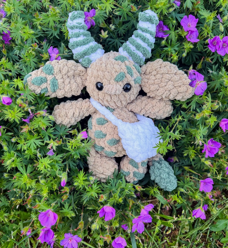 Peut inclure: Une peluche crochet&eacute;e &agrave; la main au design fantaisiste. Le jouet pr&eacute;sente un corps beige avec des taches vertes, de grandes oreilles et des cornes ray&eacute;es. Il a une sacoche blanche et est entour&eacute; de feuillage vert et de fleurs violettes.