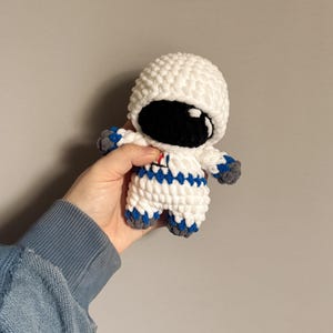 May include: A crocheted astronaut toy with a white helmet, black face, and blue and white striped suit.