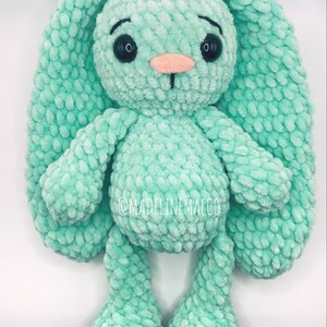 May include: A crocheted plush bunny toy in a light green color with black eyes and a pink nose. The bunny has long floppy ears and is standing on a white background.
