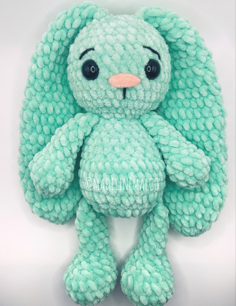 May include: A crocheted plush bunny toy in a light green color with black eyes and a pink nose. The bunny has long floppy ears and is standing on a white background.