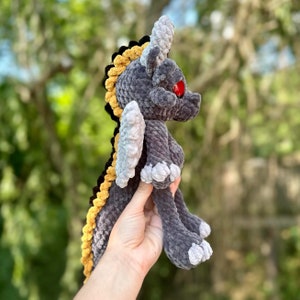 May include: A gray and yellow crocheted dragon plush toy with red eyes. The dragon has a long tail and wings.