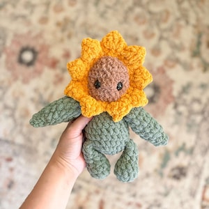K&ouml;nnte beinhalten: Ein geh&auml;keltes Pl&uuml;schtier in Form einer Sonnenblume mit gr&uuml;nem K&ouml;rper und gelbem und braunem Gesicht. Das Spielzeug wird in einer Hand gehalten.