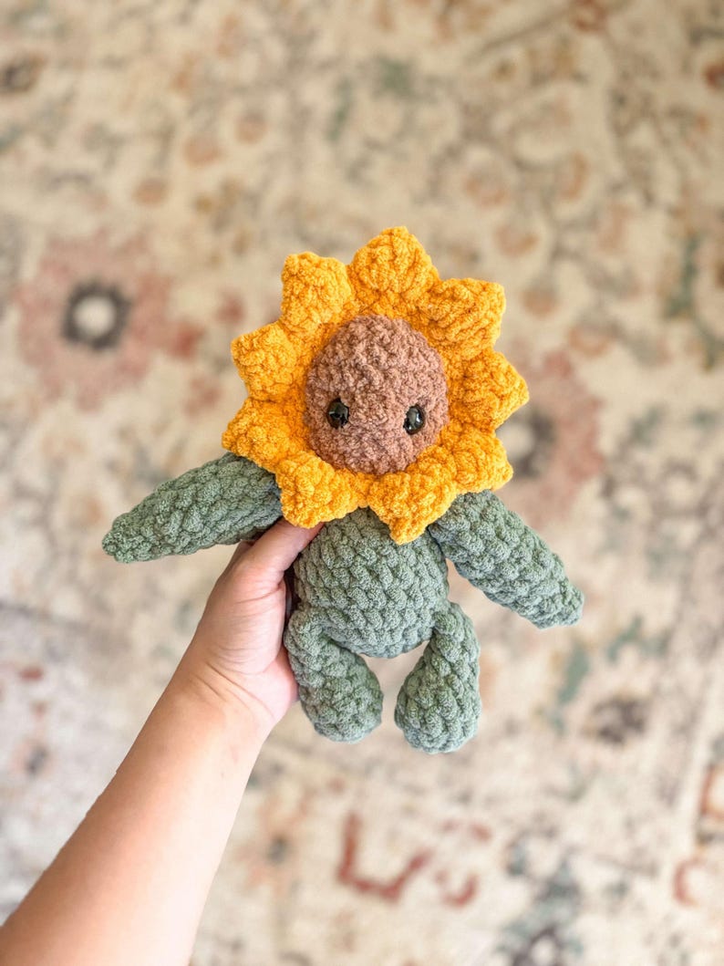K&ouml;nnte beinhalten: Ein geh&auml;keltes Pl&uuml;schtier in Form einer Sonnenblume mit gr&uuml;nem K&ouml;rper und gelbem und braunem Gesicht. Das Spielzeug wird in einer Hand gehalten.