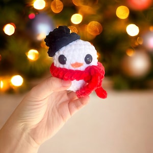 May include: A small white crocheted snowman with a black hat and a red scarf. The snowman is being held in a hand against a blurry background of Christmas lights.