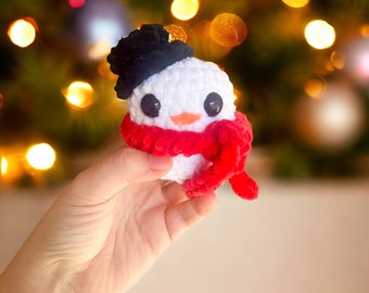 May include: A small white crocheted snowman with a black hat and a red scarf. The snowman is being held in a hand against a blurry background of Christmas lights.