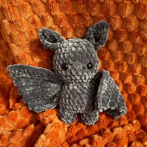 Könnte beinhalten: Ein dunkelgraues Häkel-Fledermaus-Stofftier aus weichem, strukturiertem Garn. Die Fledermaus hat zwei spitze Ohren, zwei kleine schwarze Kunststoffaugen und breite, detaillierte Flügel, die von ihrem runden Körper abstehen. Die Oberfläche wirkt flauschig.