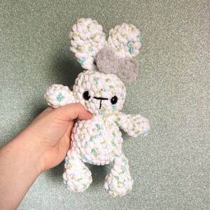 May include: A handmade white and blue crochet bunny with a gray bow and black eyes. The plush toy has a soft, textured appearance, with a cute, whimsical design. The bunny's ears are large and floppy, and its body is small and round.