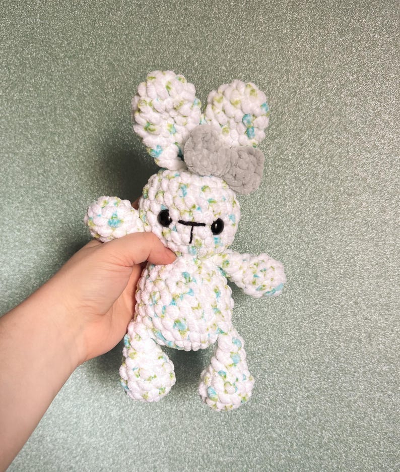 May include: A handmade white and blue crochet bunny with a gray bow and black eyes. The plush toy has a soft, textured appearance, with a cute, whimsical design. The bunny's ears are large and floppy, and its body is small and round.