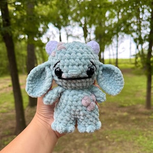 May include: A light blue crocheted plush toy with a flower on its chest. The toy has large eyes and a small mouth. It has two small horns on its head.