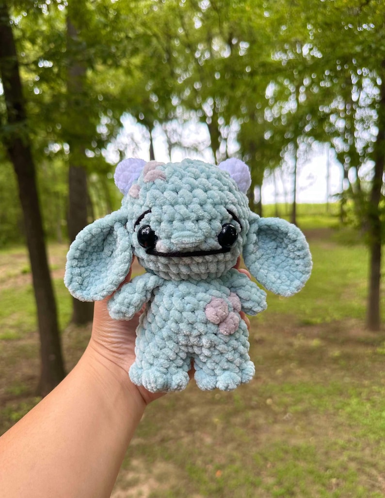 May include: A light blue crocheted plush toy with a flower on its chest. The toy has large eyes and a small mouth. It has two small horns on its head.